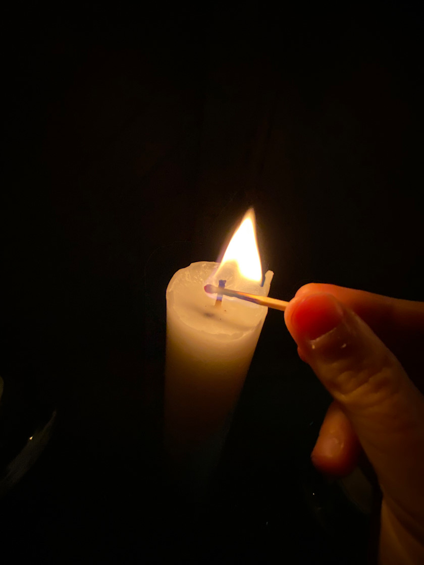 En person tänder ett ljus i ett mörkt rum. Man ser bara personens fingrar som håller i en tändsticka och lågan från tändstickan som tänder ljuset. [foto: Carlota "Carro" Bocio Duff]
